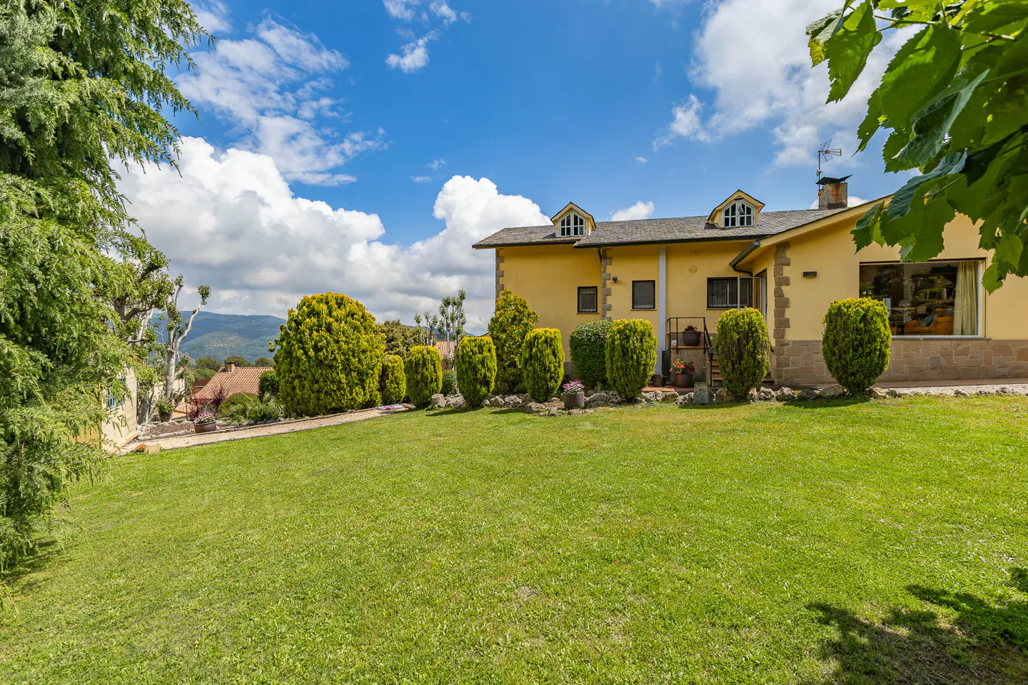 A yellow house with dormer windows sits on a green lawn under a blue sky with white clouds.