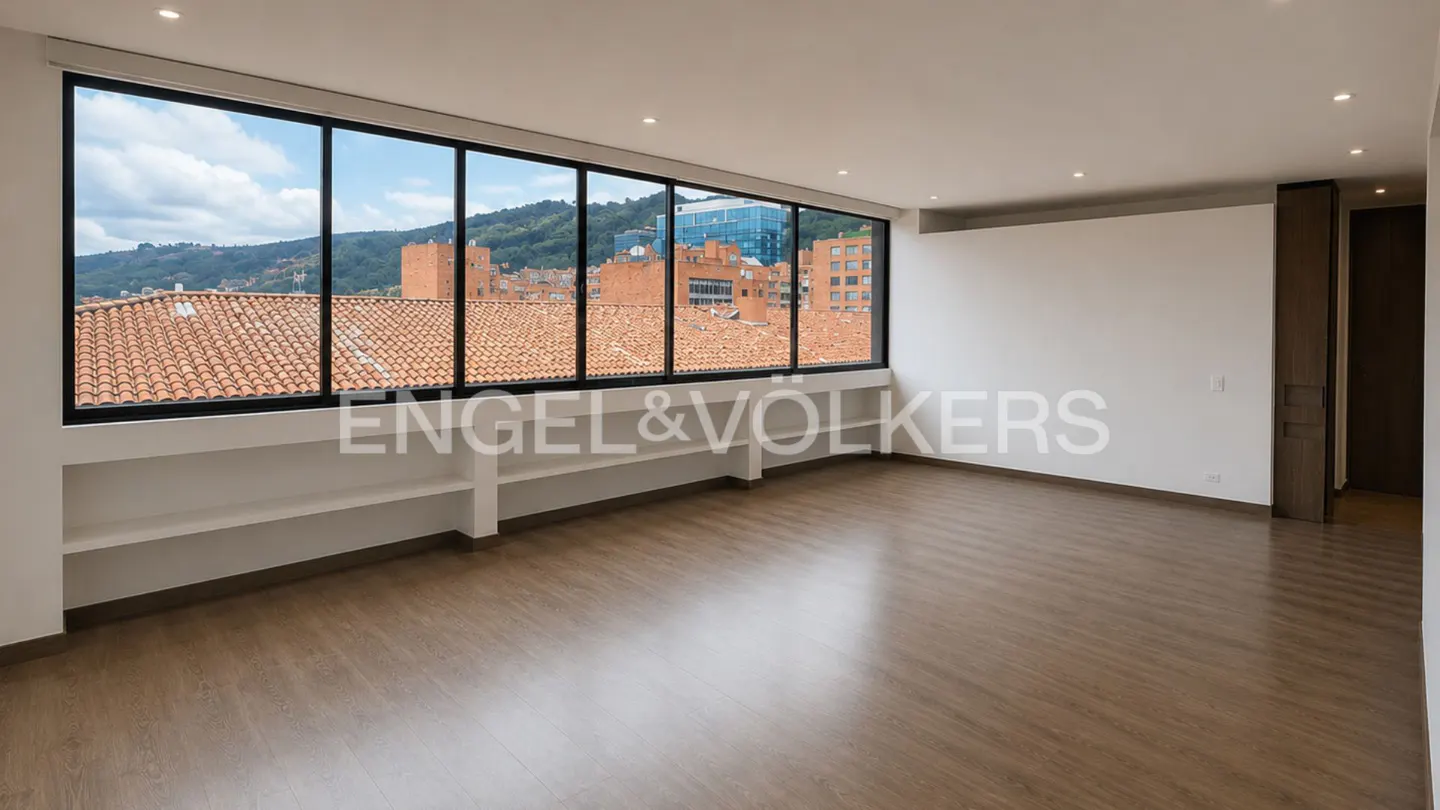 Empty room with light wood floors, white walls, and a large window overlooking city rooftops and distant mountains.