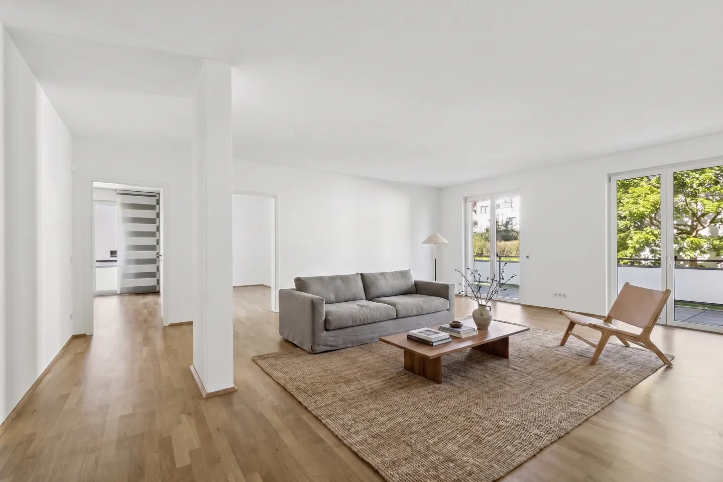 Bright, modern living room with hardwood floors, a gray sofa, a wood coffee table, and a tan woven rug. Balcony doors let in natural light.