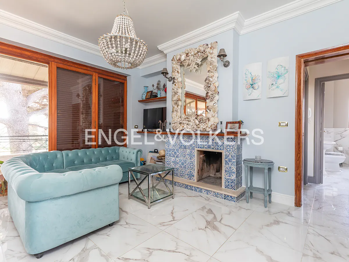 Living room with a blue sofa, a fireplace with a seashell mirror, and a chandelier. The floor is white marble.
