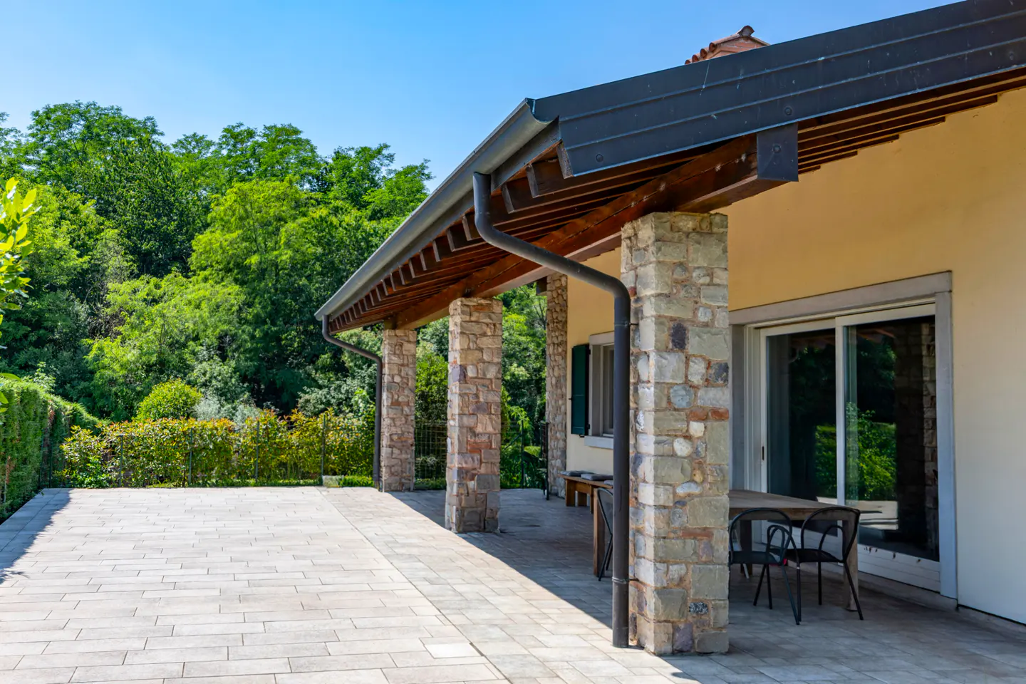 A stone-pillared patio with a wooden roof, outdoor dining set, and a view of lush green trees under a clear blue sky.