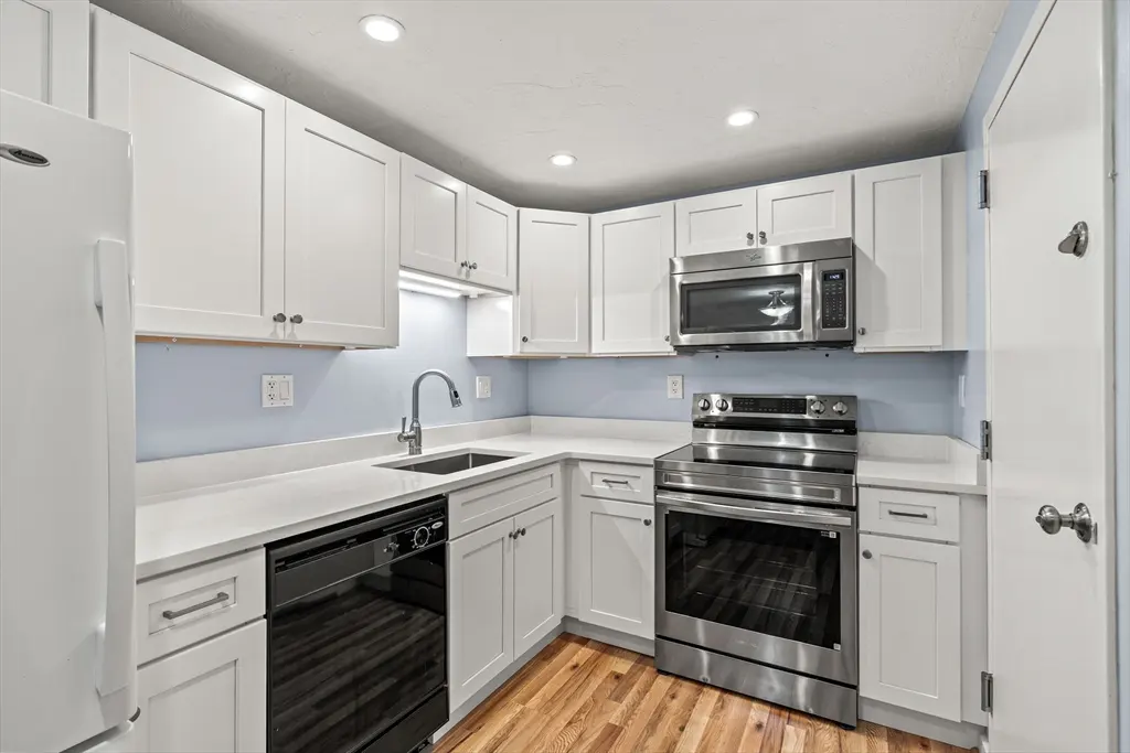 A bright kitchen with white cabinets, stainless steel appliances, and light blue walls. A black dishwasher is next to the sink. The floor is wood.