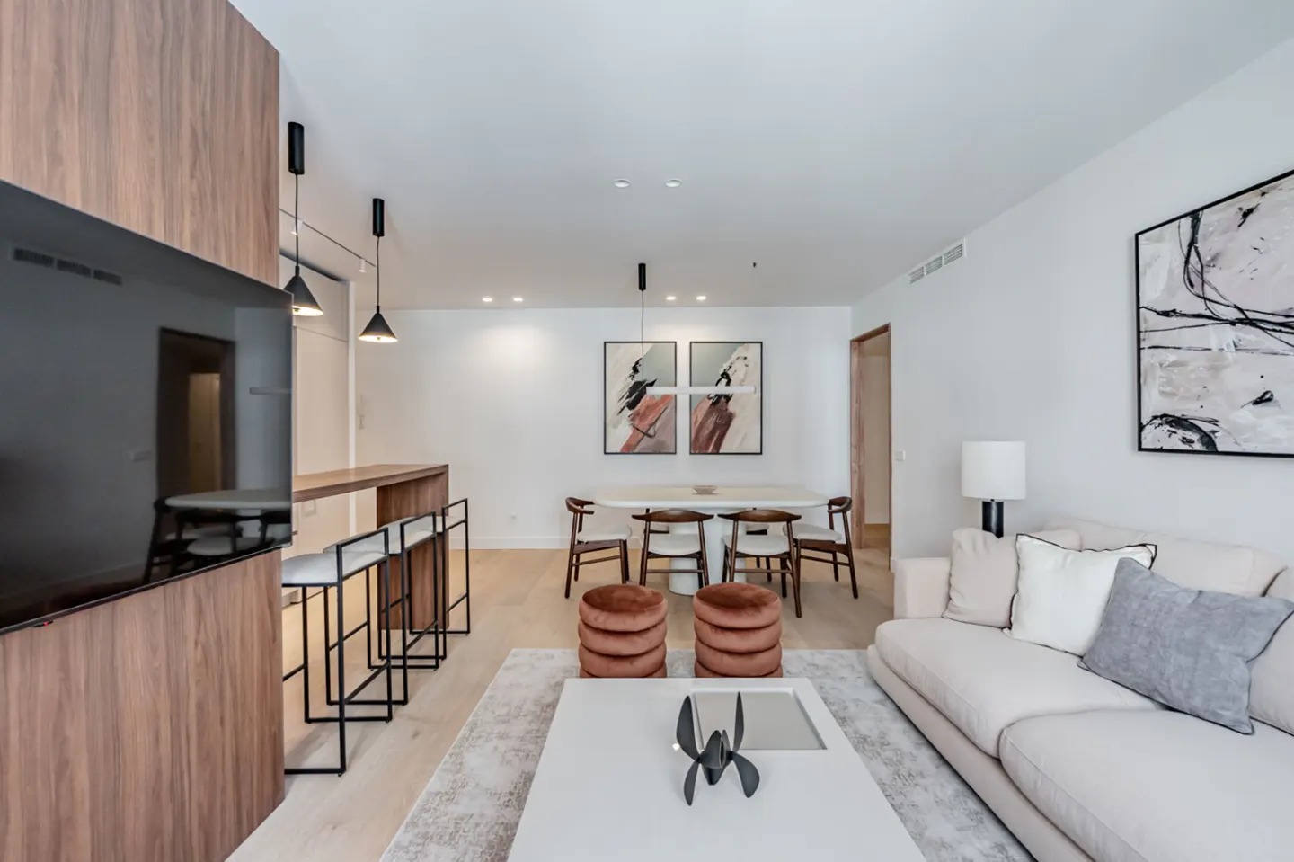 Modern living room with white walls, light wood floors, and a large TV. A white sofa, coffee table, and dining table are visible.