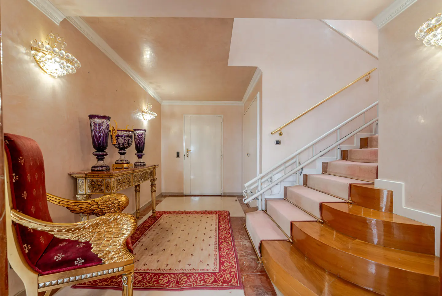 Elegant foyer with pink walls, ornate gold chair, console table with purple vases, and a staircase with pink carpet.