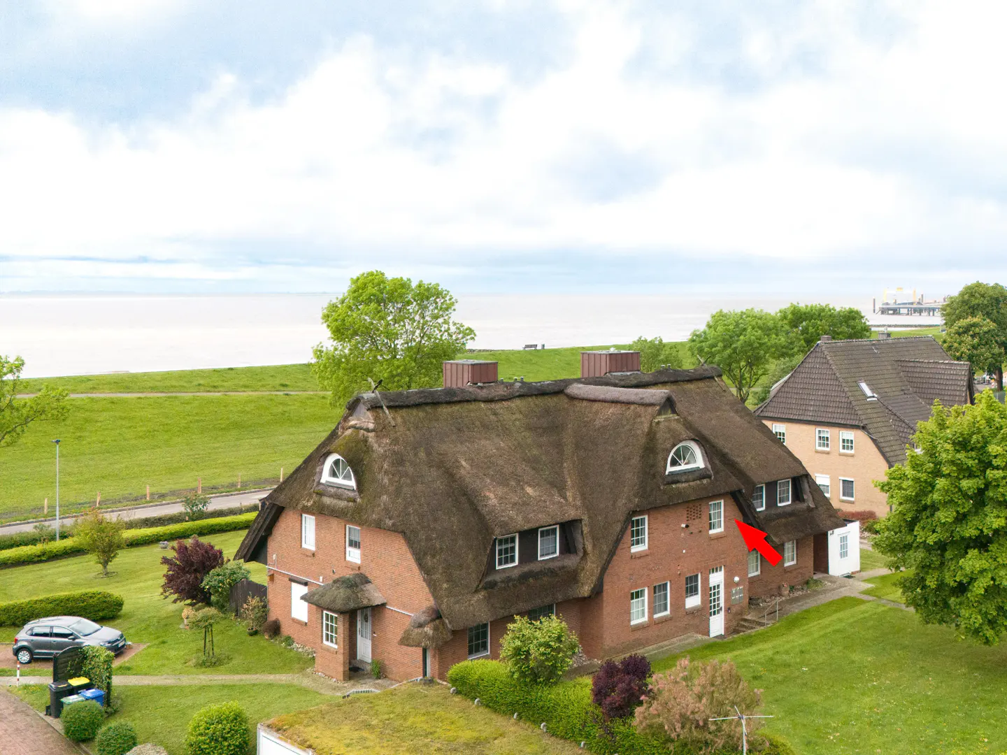 Aerial view of a red brick house with a thatched roof near a grassy field and the sea, with a red arrow pointing to a window.