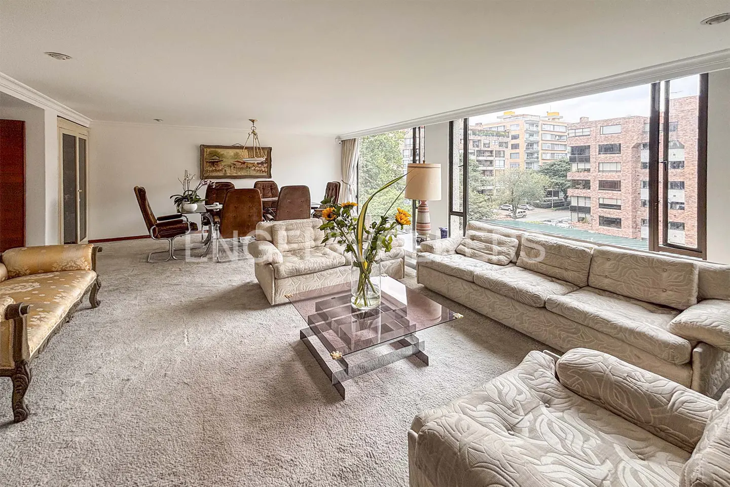 Spacious living room with beige sofas, glass coffee table, and dining area. Large windows offer city views.