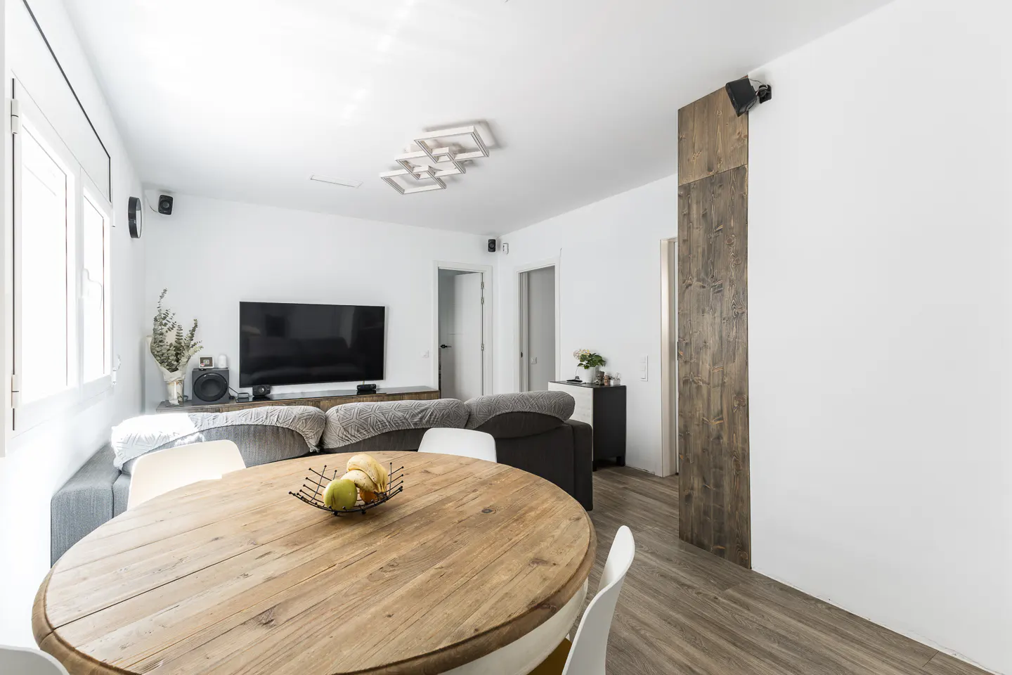 Bright living room with a round wooden table, white chairs, and a fruit bowl. A gray sofa, TV, and speakers are in the background.