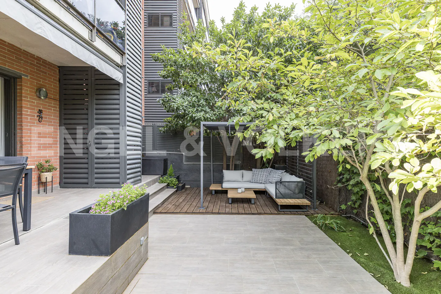 A modern patio with gray tile, a gray planter with flowers, and a gray outdoor sofa under a pergola surrounded by lush green trees.