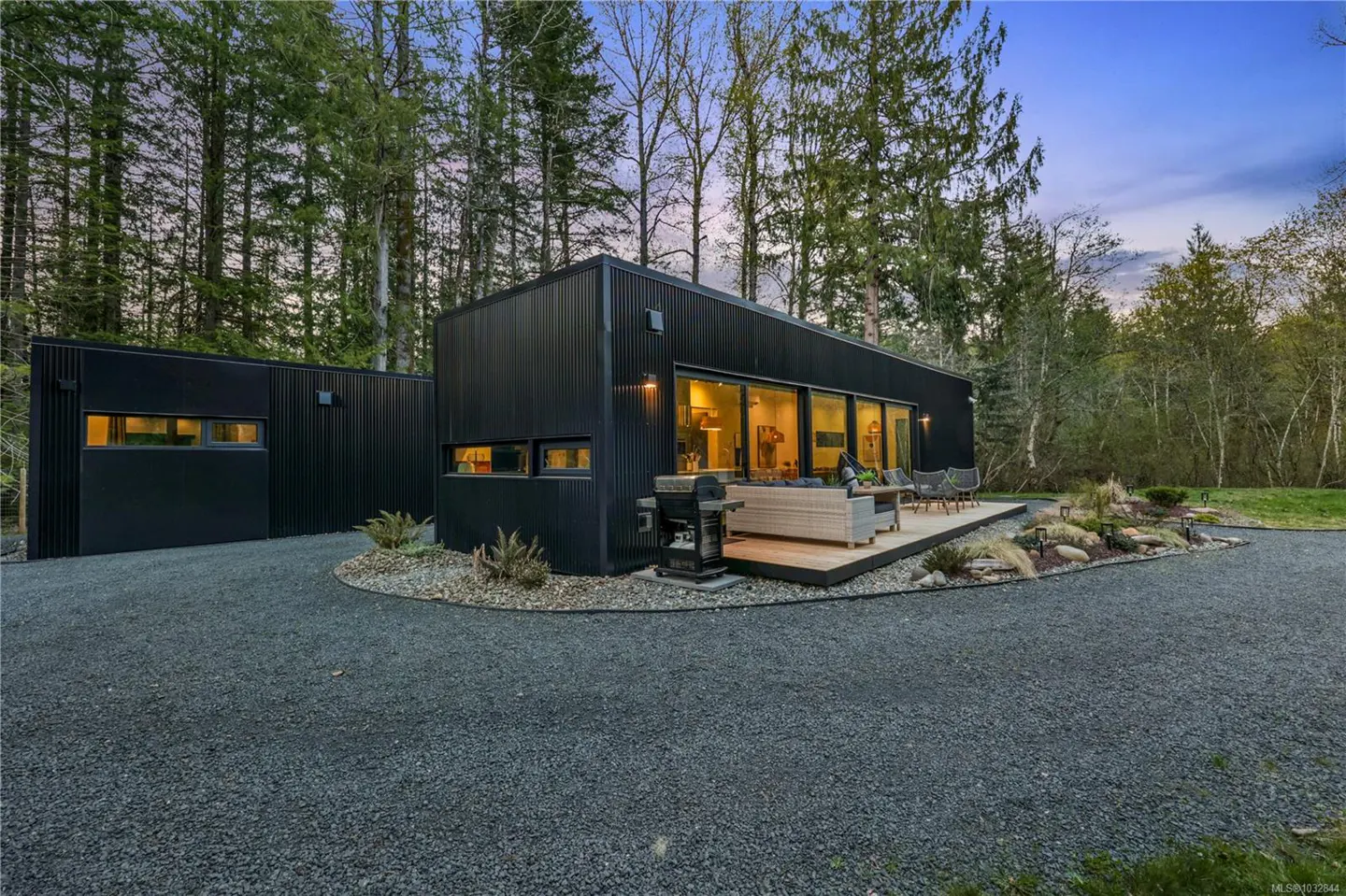Modern black home with a gravel driveway and a wooden deck. Large windows reflect warm light. Trees surround the property.