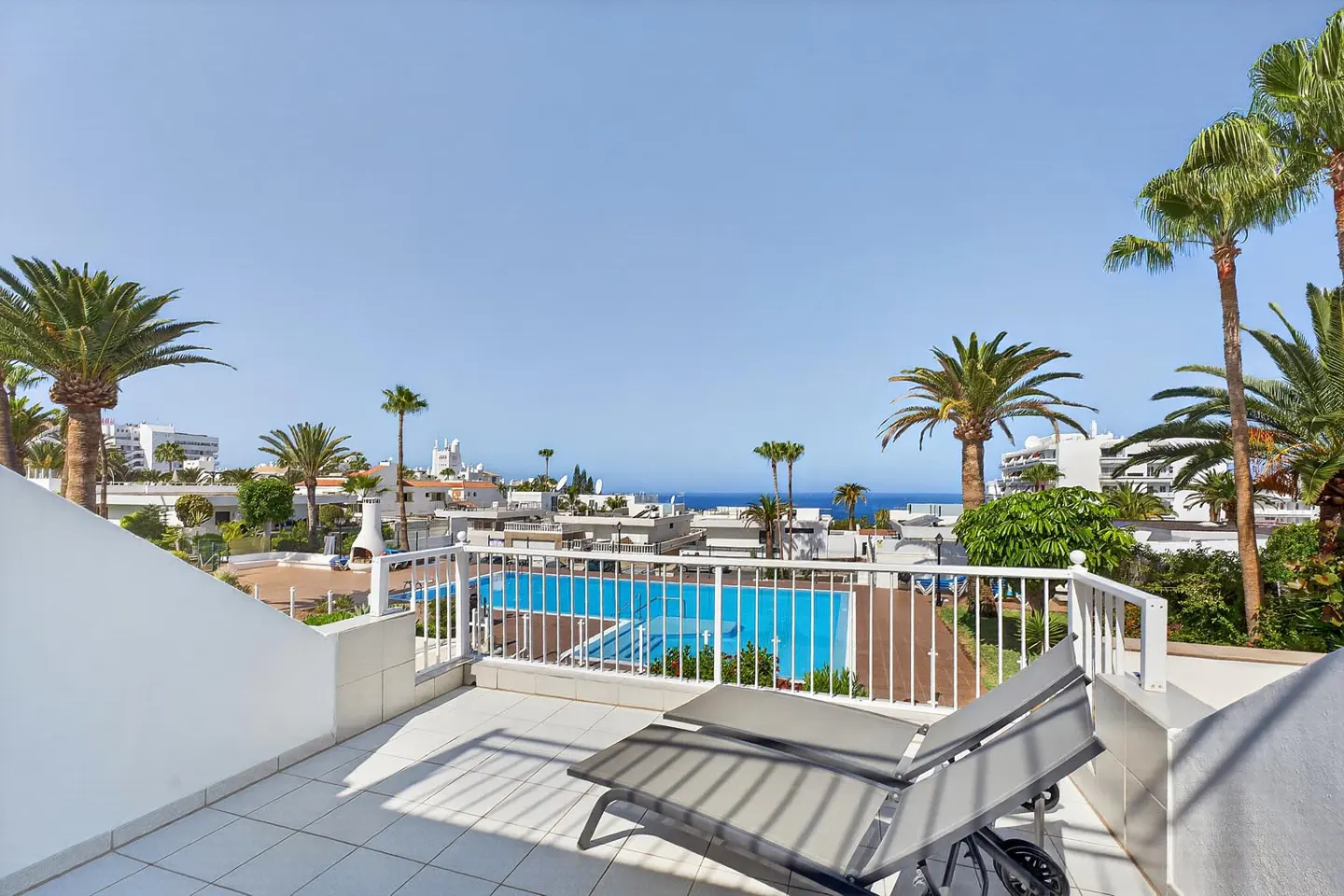 Sunny balcony with two grey lounge chairs overlooking a blue swimming pool, palm trees, and the ocean in the distance.