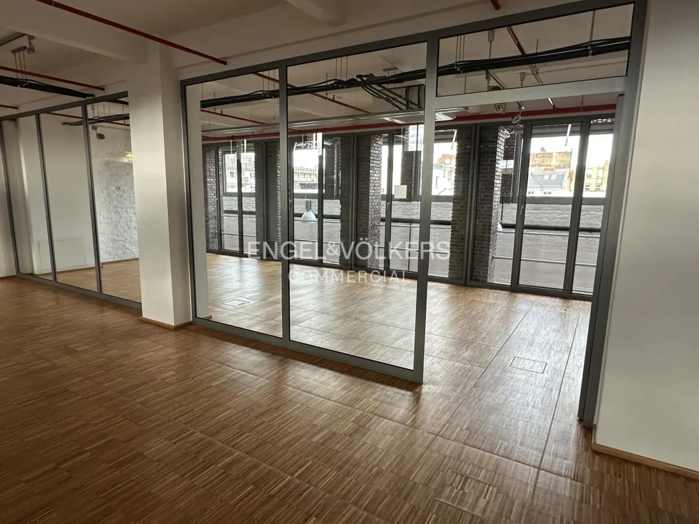 Empty office space with wood floors, glass-walled rooms, white walls, and exposed brick columns. The Engel & Völkers logo is visible.