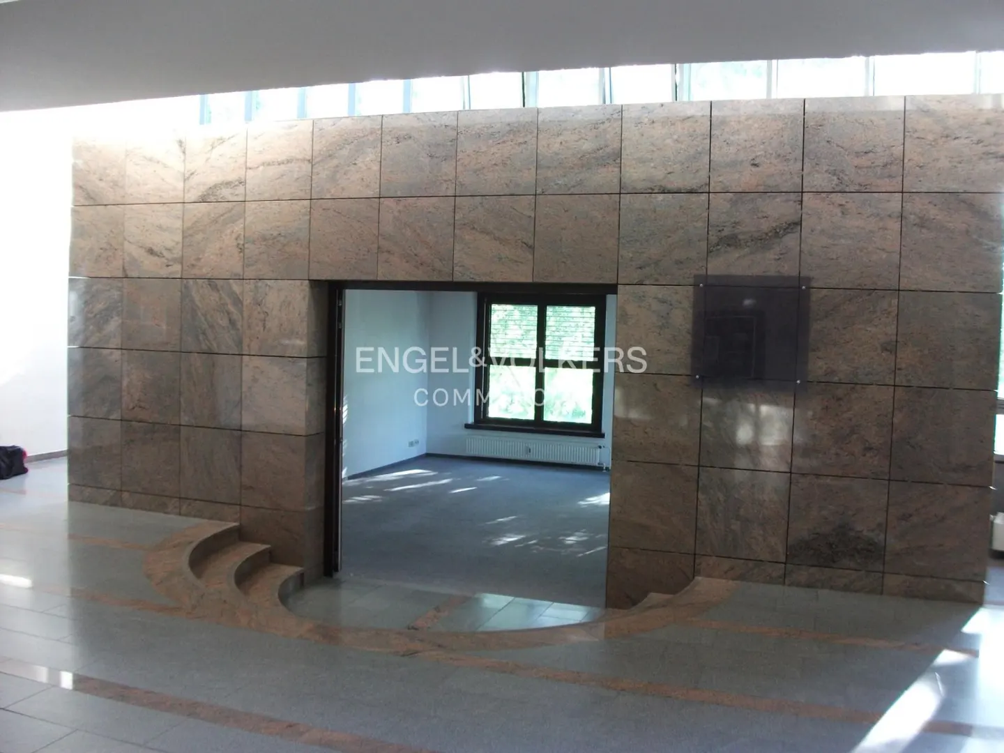 Interior view of a building with a marble wall and doorway. A room with a window is visible through the doorway. The floor is gray with marble accents.