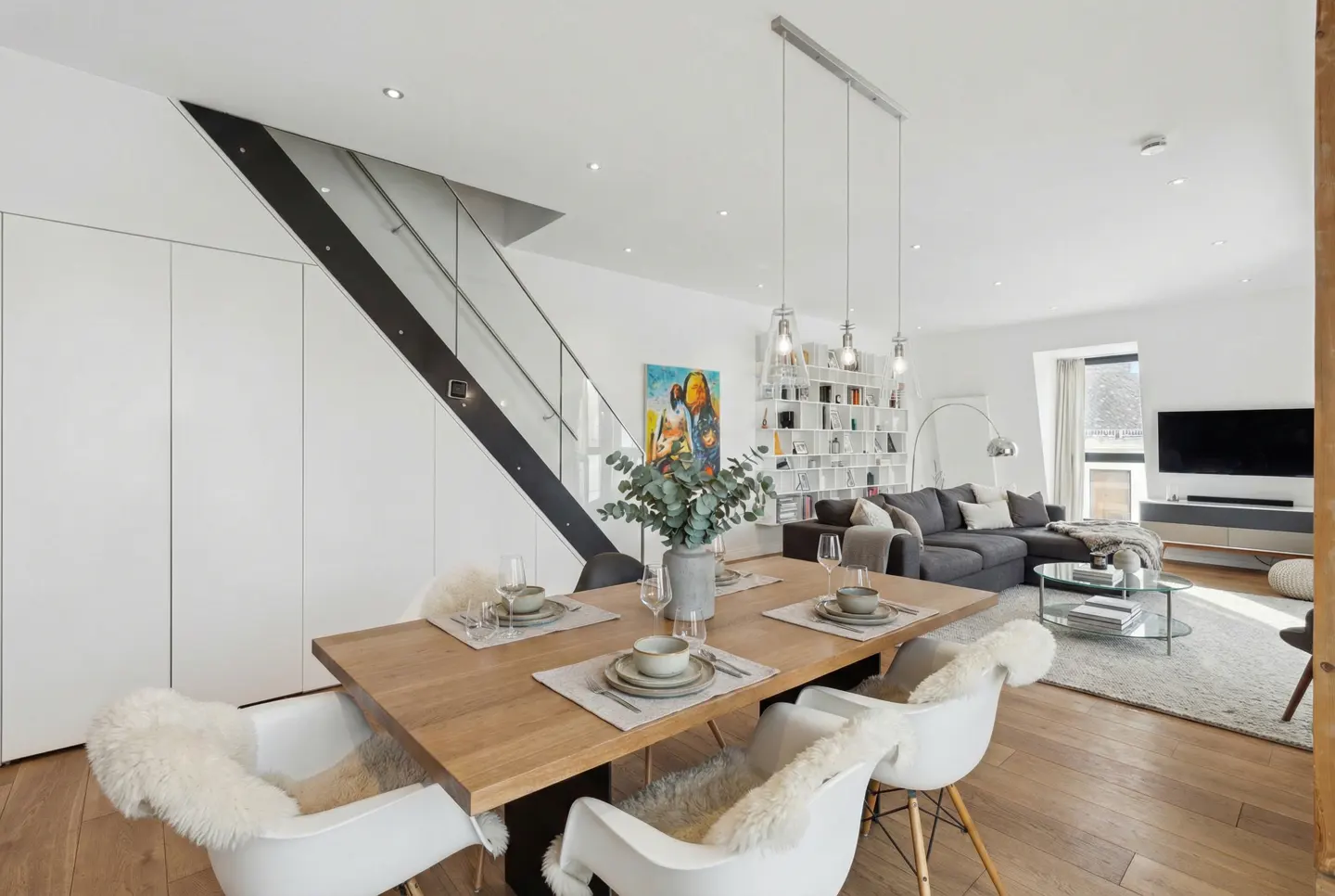 Bright, modern dining and living space. A wood table with place settings sits in front of a gray sofa, TV, and white shelving. Stairs are on the left.