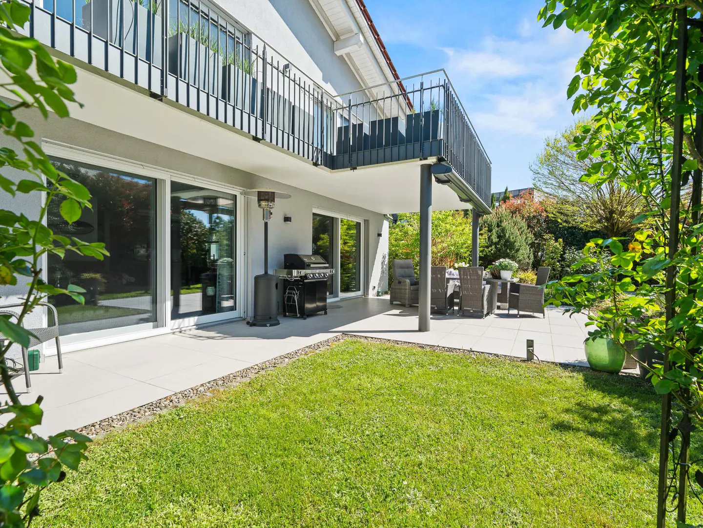 A modern house with a patio, grill, and outdoor seating overlooks a green lawn and garden.