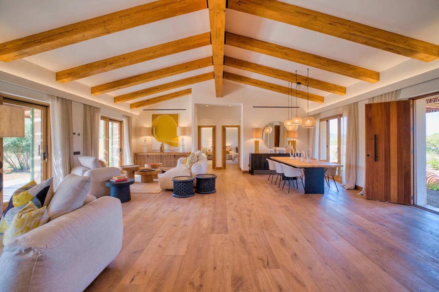 Open-concept living space with wood floors, exposed beams, and neutral furniture. Dining table with pendant lights. Large windows and doors.
