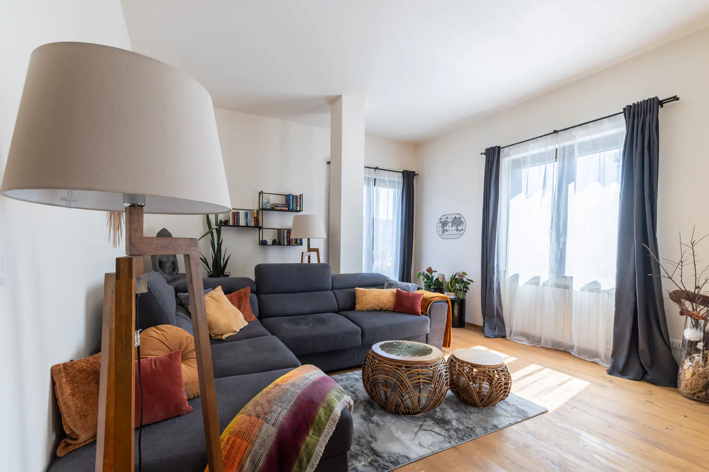 Bright living room with gray sectional sofa, colorful pillows, and a large floor lamp. Wicker tables sit on a gray rug. Sunlight streams through sheer curtains.