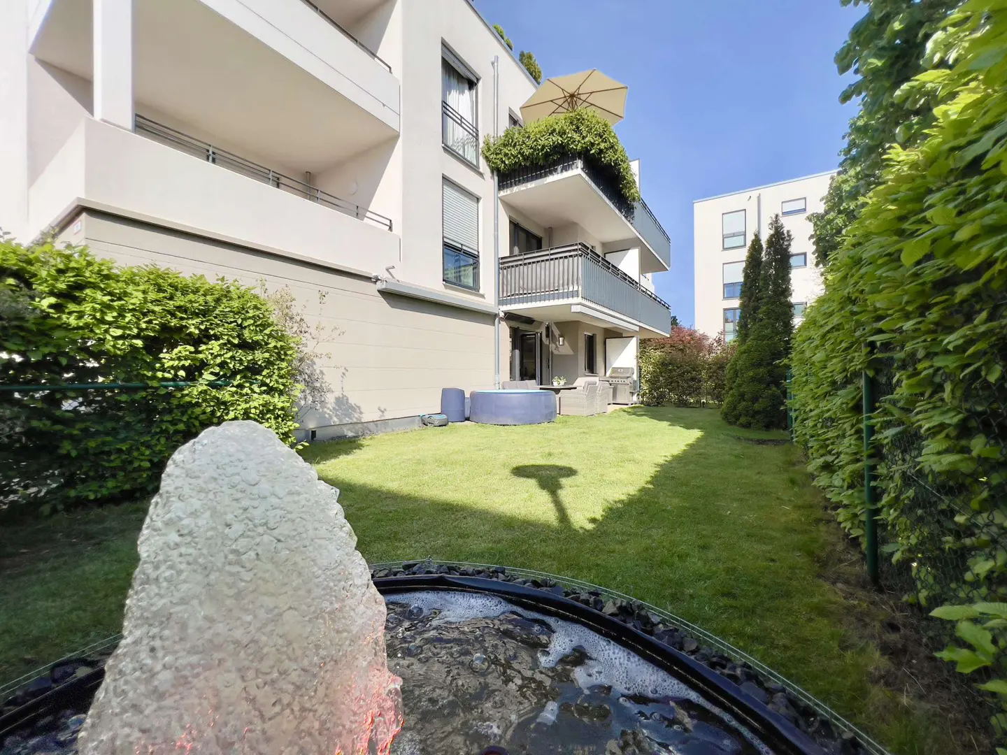 A modern apartment building with balconies overlooks a green lawn and a fountain in the foreground.