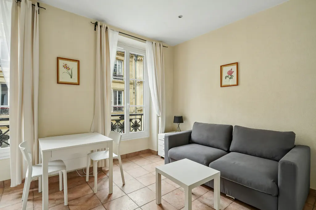 A cozy apartment interior with a gray sofa, white table and chairs, and a window view of a Parisian building.