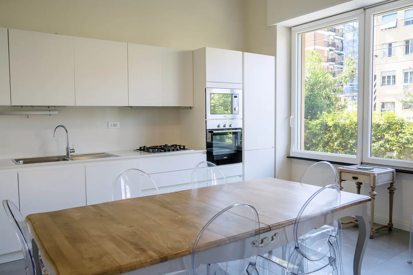 Bright kitchen with white cabinets, stainless steel appliances, and a wood table with clear chairs. Large window with city view.