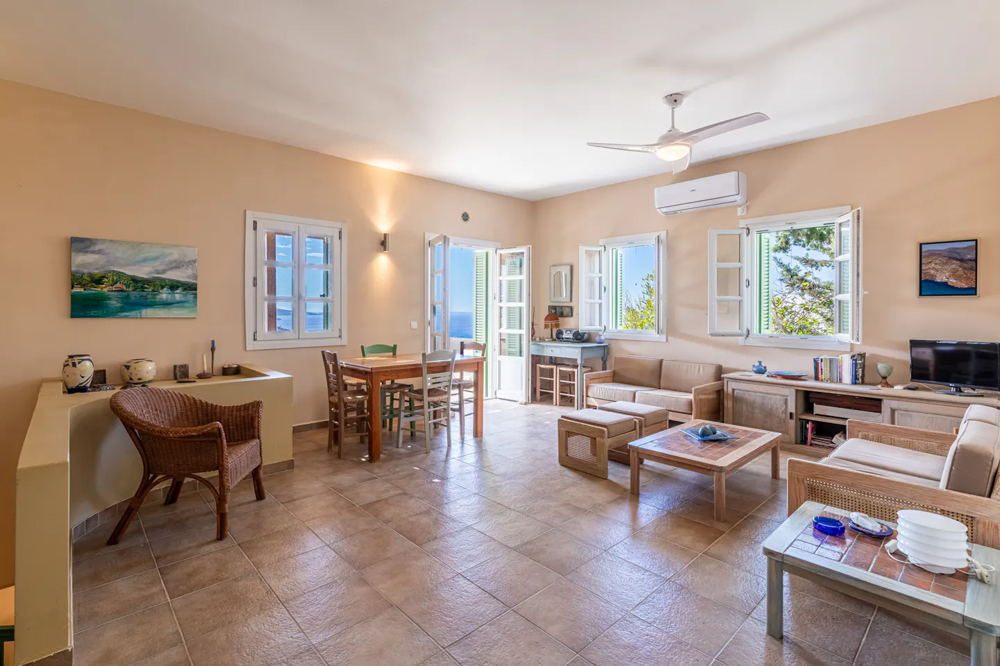 Bright living room with tile floor, beige walls, and white trim. Furniture includes sofas, tables, and a dining set. Windows offer ocean views.