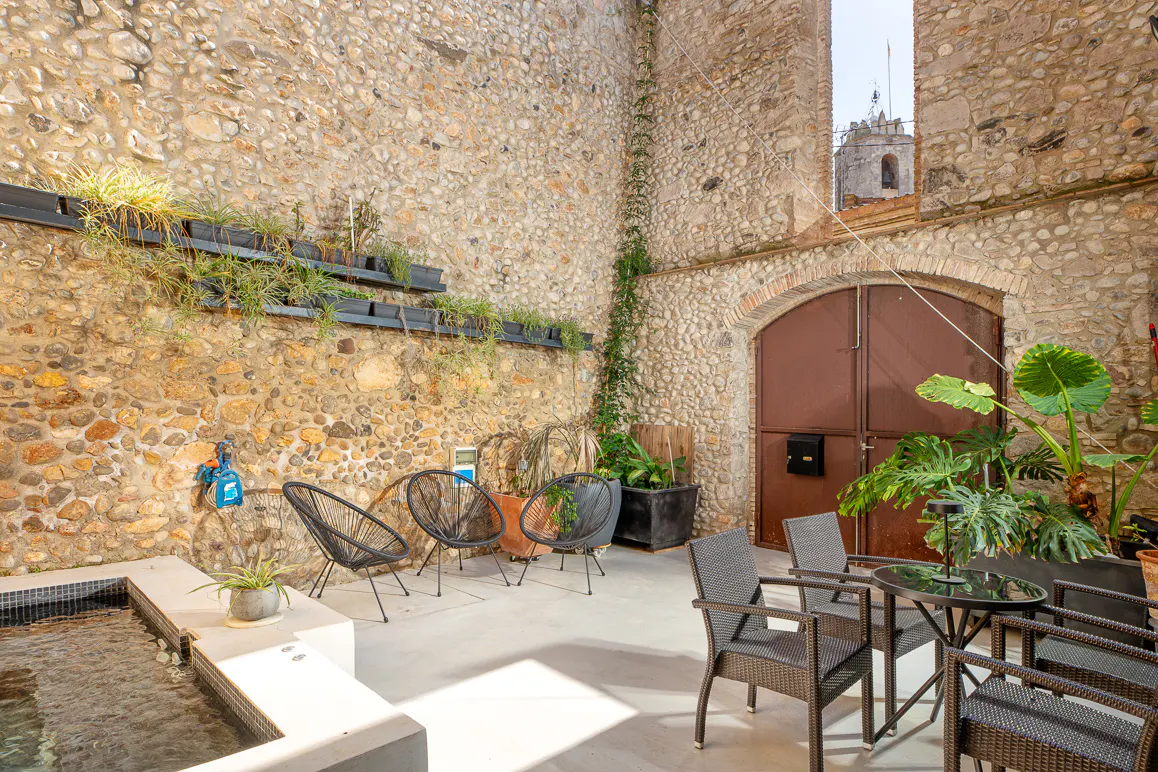 Outdoor patio with stone walls, plants, and seating. A small pool is visible on the left. A church tower is seen in the background.