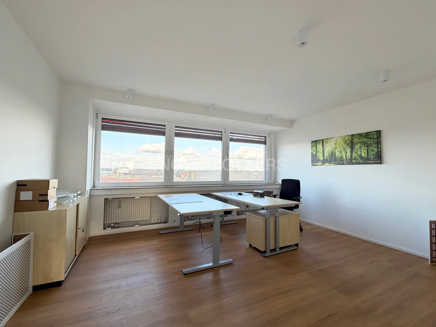 Bright office space with wood floors, white walls, and large windows offering a city view. Two desks and a black office chair are present.