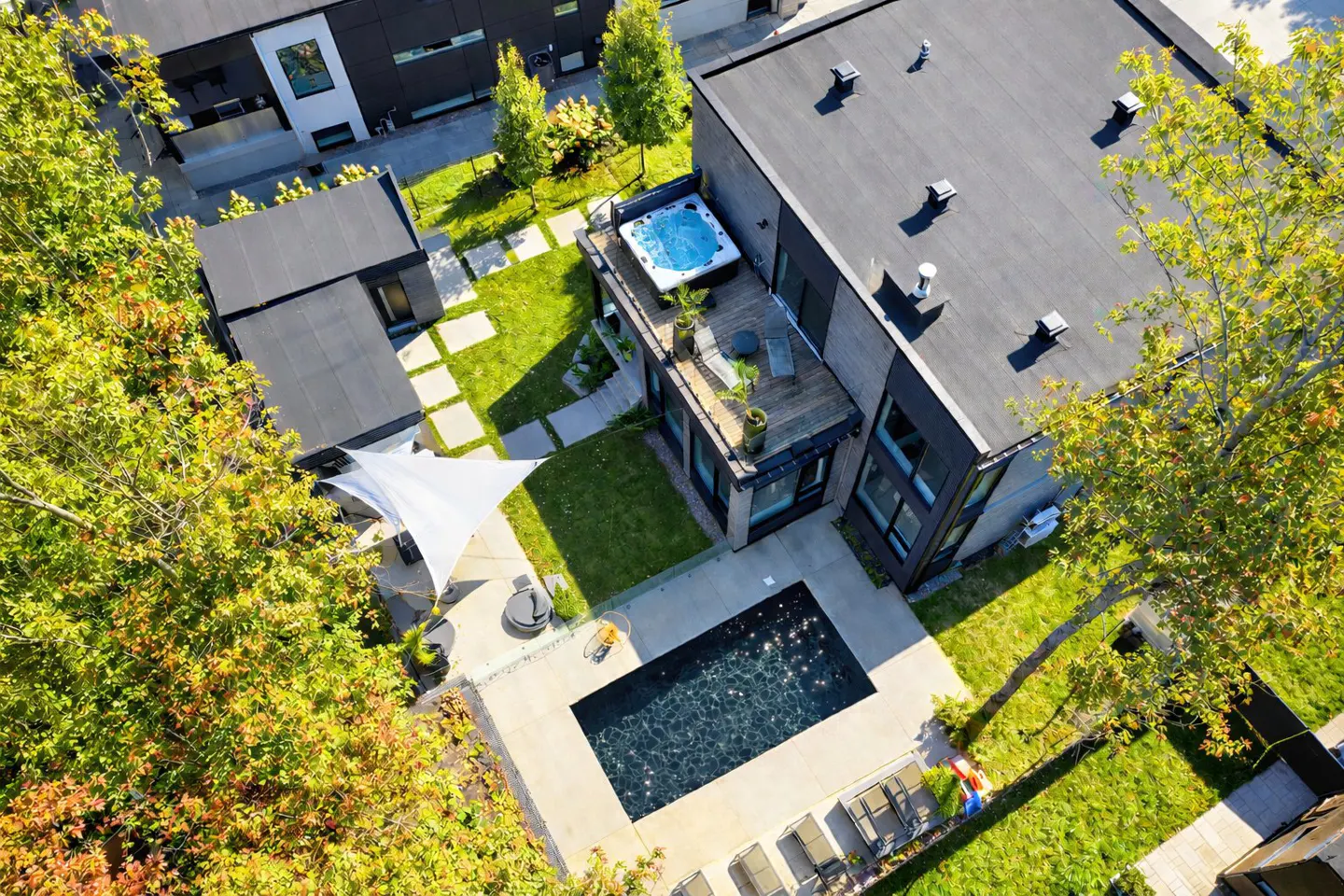 Aerial view of a modern home with a pool, hot tub, and lush green landscaping. The house has a flat roof and large windows.