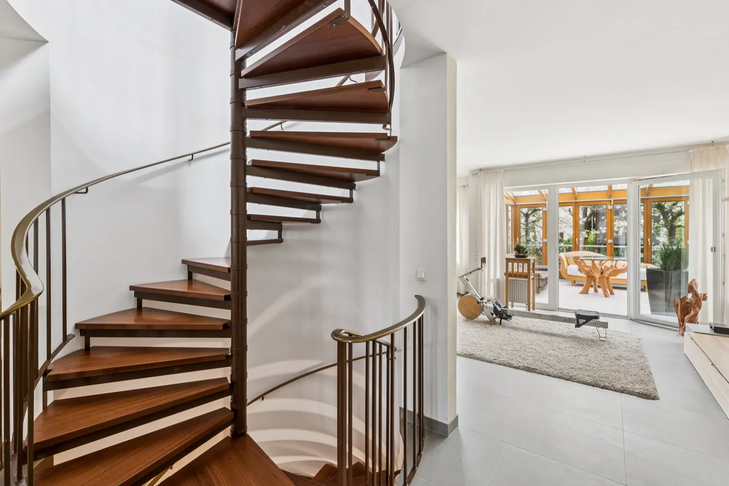 Interior view of a home featuring a wooden spiral staircase and a bright living area with exercise equipment.