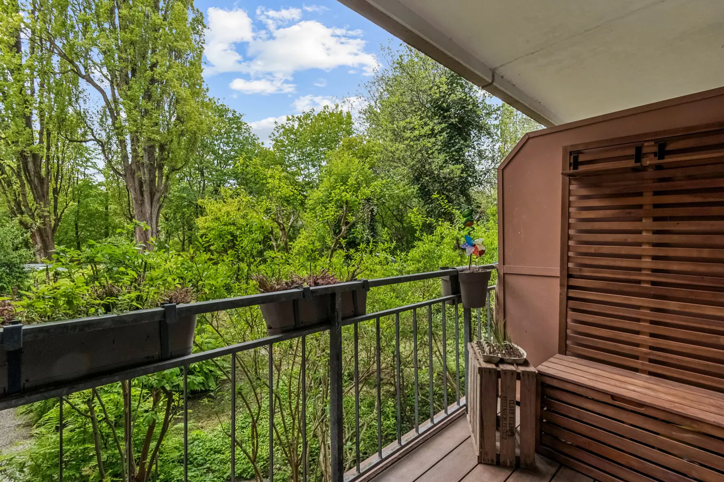 Balcony view with metal railing, potted plants, and wooden bench. Lush green trees fill the background under a blue sky.