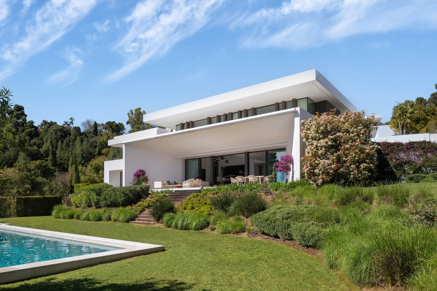 Modern white house with a flat roof, large windows, and a covered patio overlooking a green lawn and pool under a blue sky.