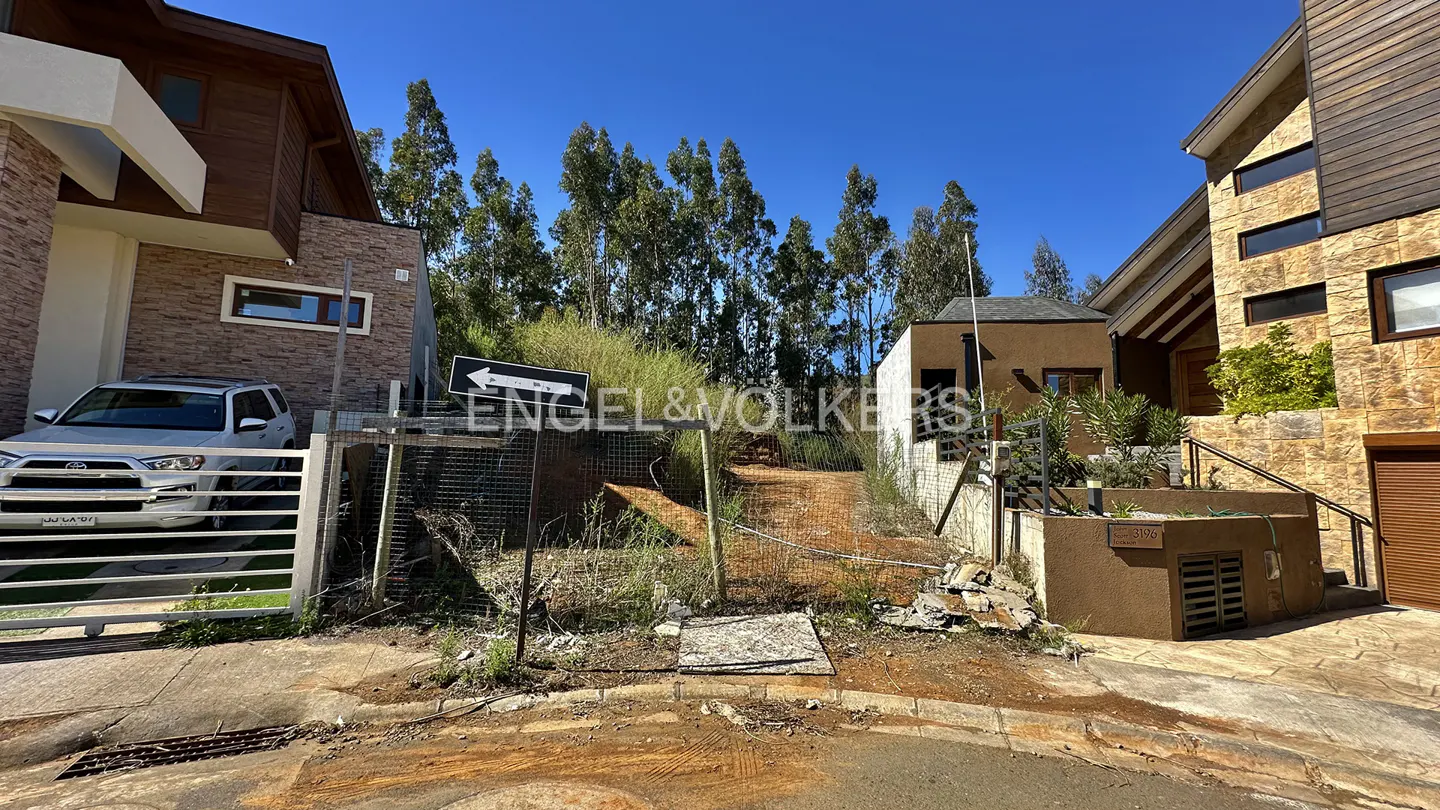 Engel & Volkers sign points to a dirt road between modern homes under a blue sky. Tall trees line the road.