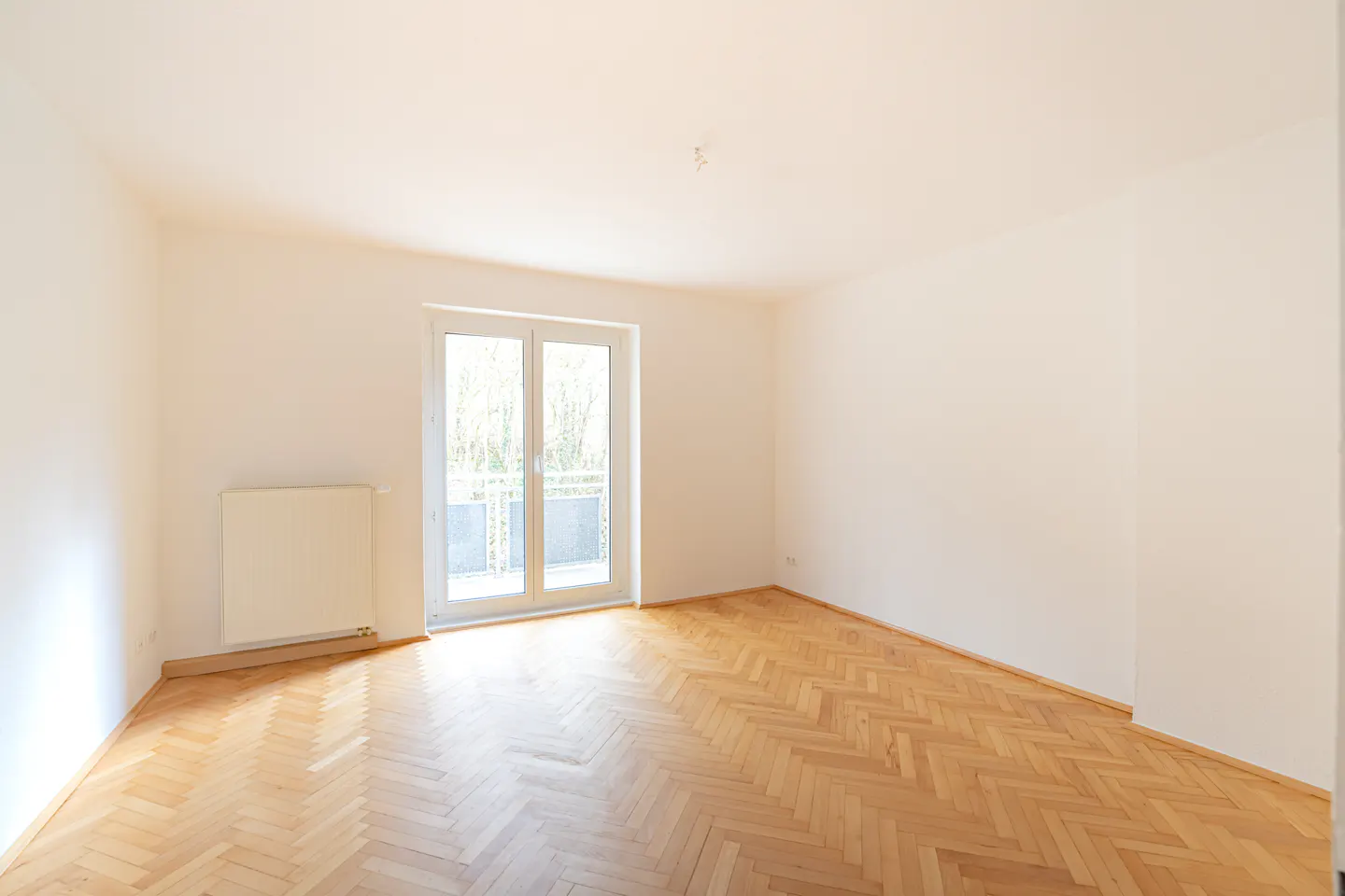 Bright, empty room with herringbone wood floors, white walls, radiator, and glass doors to a balcony.