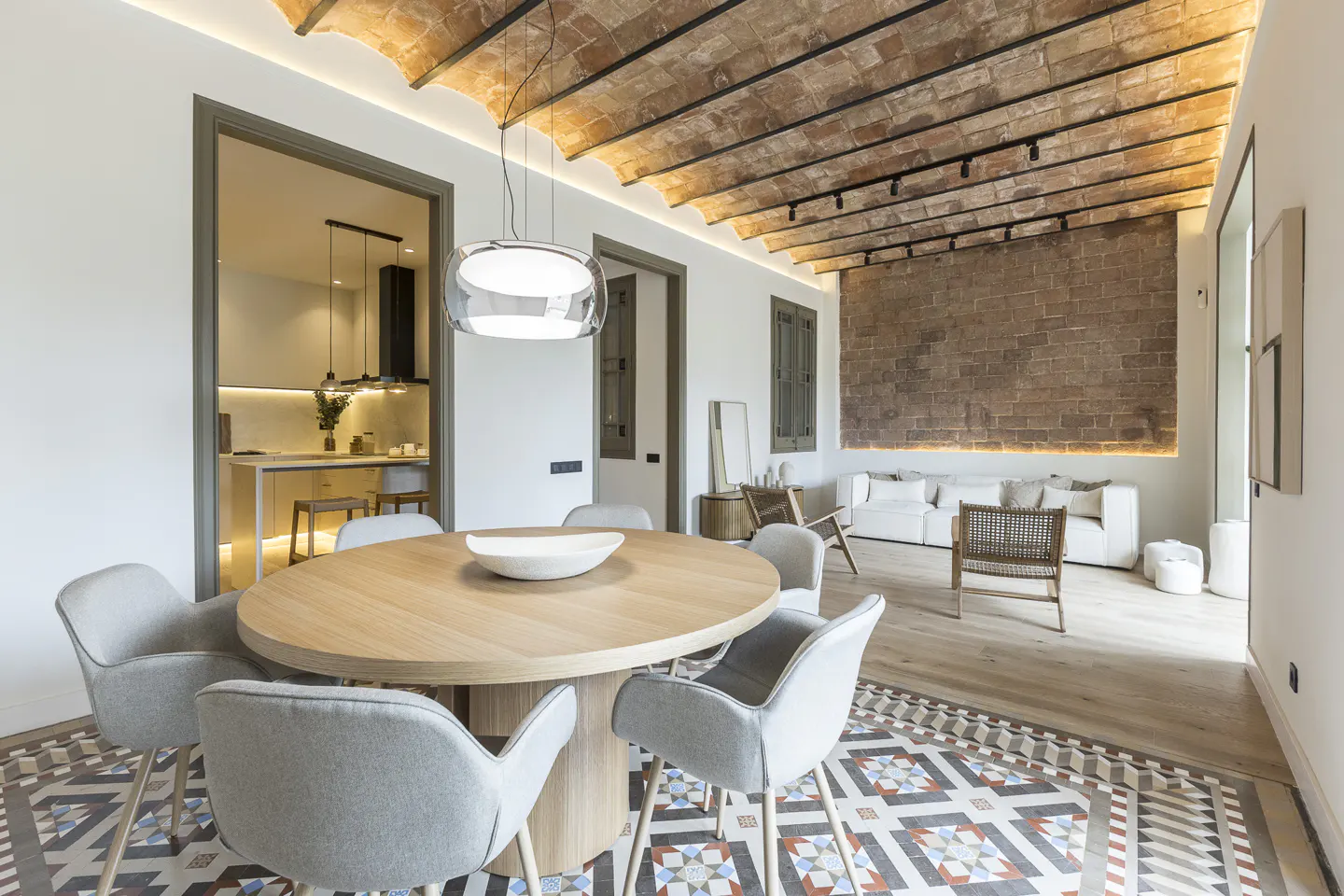 Modern dining room with a round wooden table, grey chairs, and patterned tile floors under a vaulted brick ceiling.