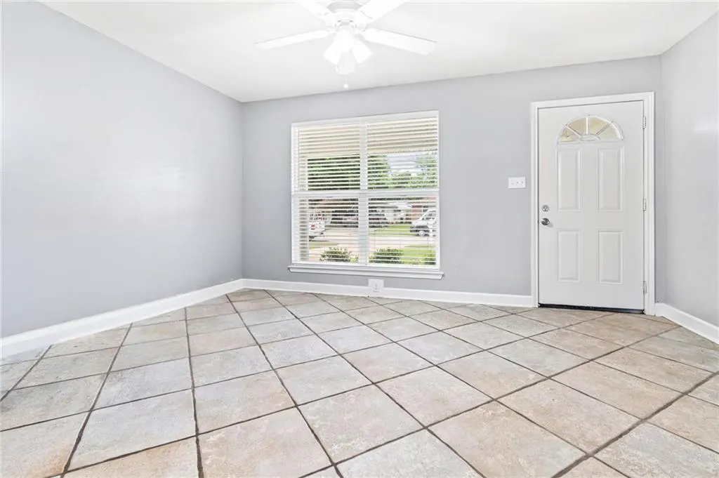 Empty room with gray walls, tile floor, and white door and trim. A window with blinds looks out onto a street. A white ceiling fan is overhead.