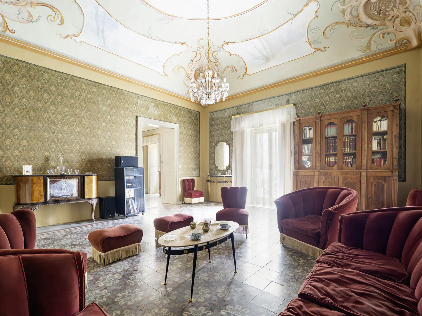 Ornate living room with red velvet seating, patterned wallpaper, and a decorative ceiling with a chandelier.