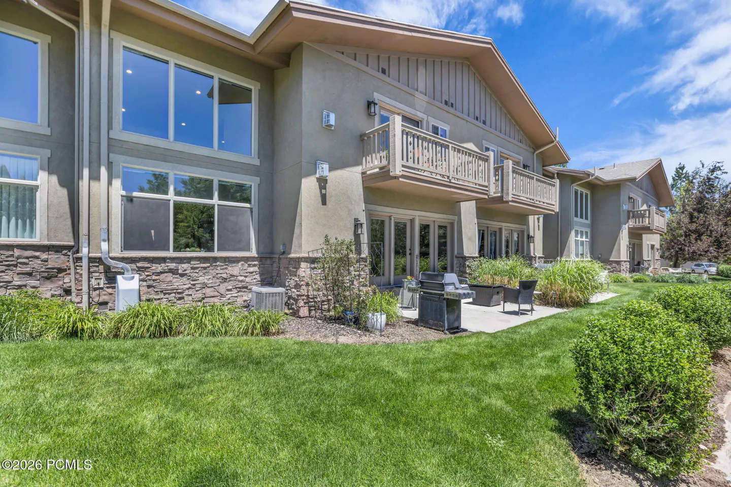 Rear view of a multi-story tan condo building with stone accents, a patio, a grill, and a green lawn under a blue sky.