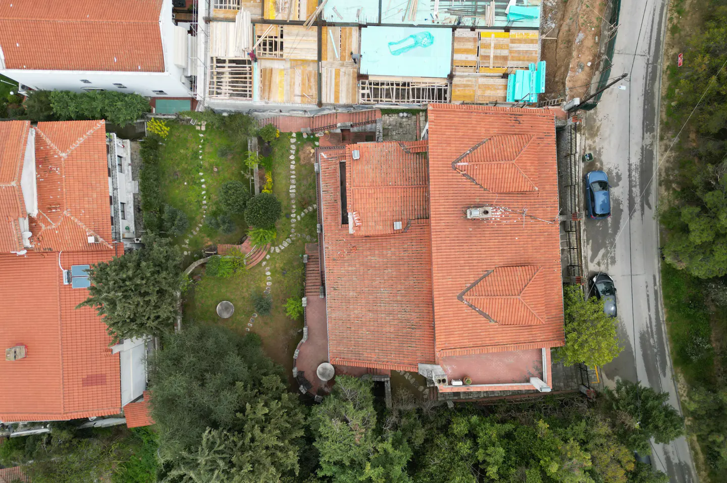 Aerial view of a house with a red tile roof, a green garden, and two cars parked on the street.