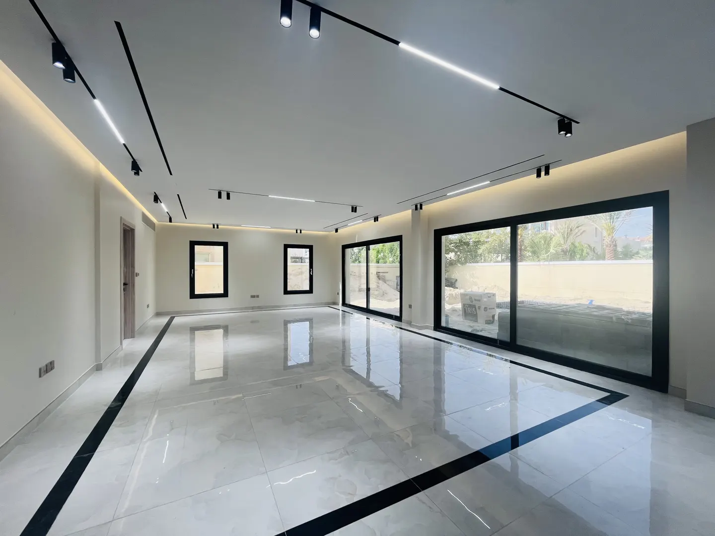 Bright, empty room with white walls, marble floors with black trim, and black-framed windows and sliding doors. Modern track lighting on the ceiling.