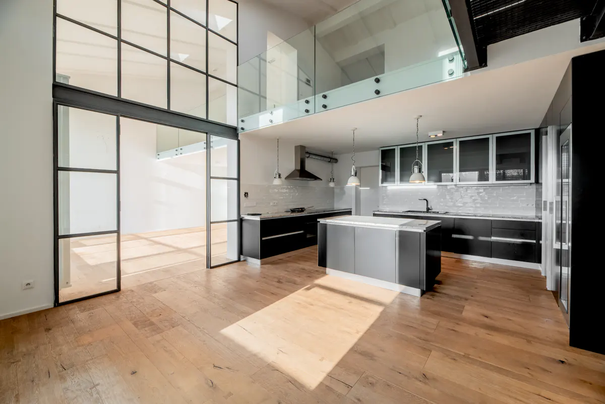 Bright, modern kitchen with wood floors, black cabinets, and a gray island. A glass-walled loft overlooks the kitchen. Black framed glass doors lead to another room.