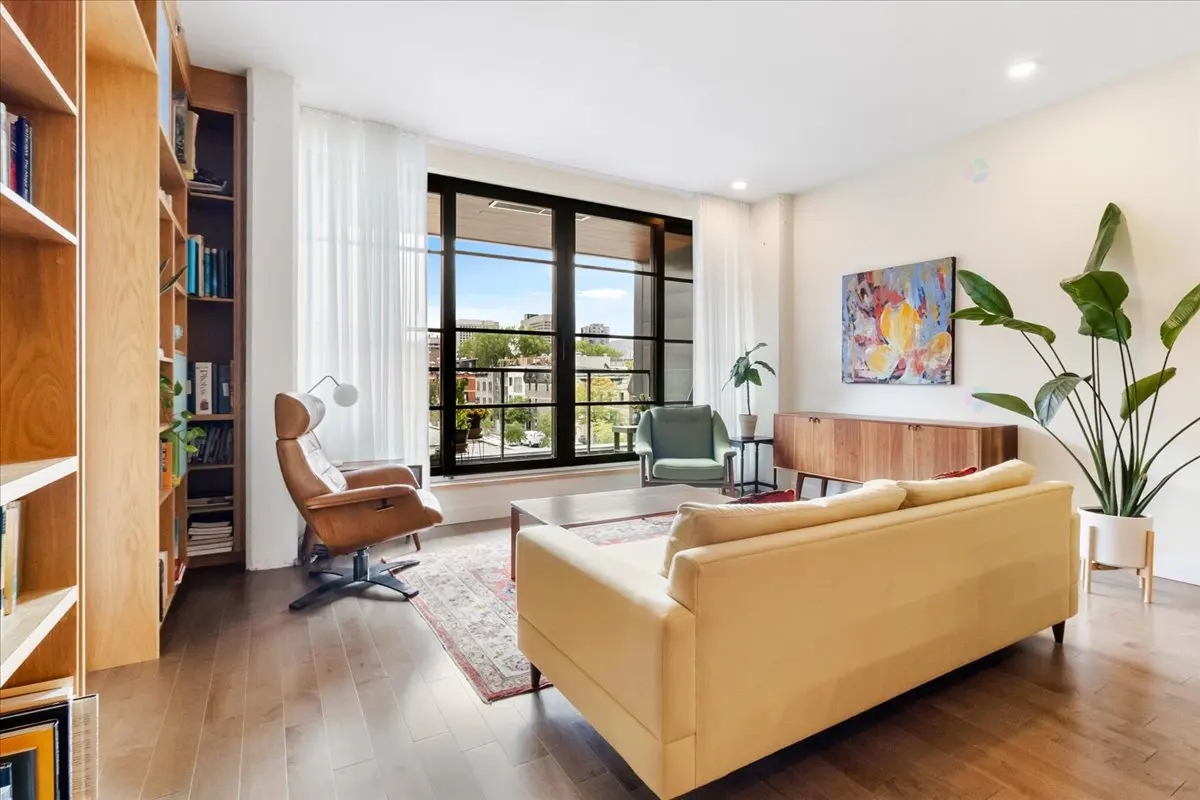 Bright living room with wood floors, a yellow sofa, and a large window with a city view. A bookshelf and artwork add character.