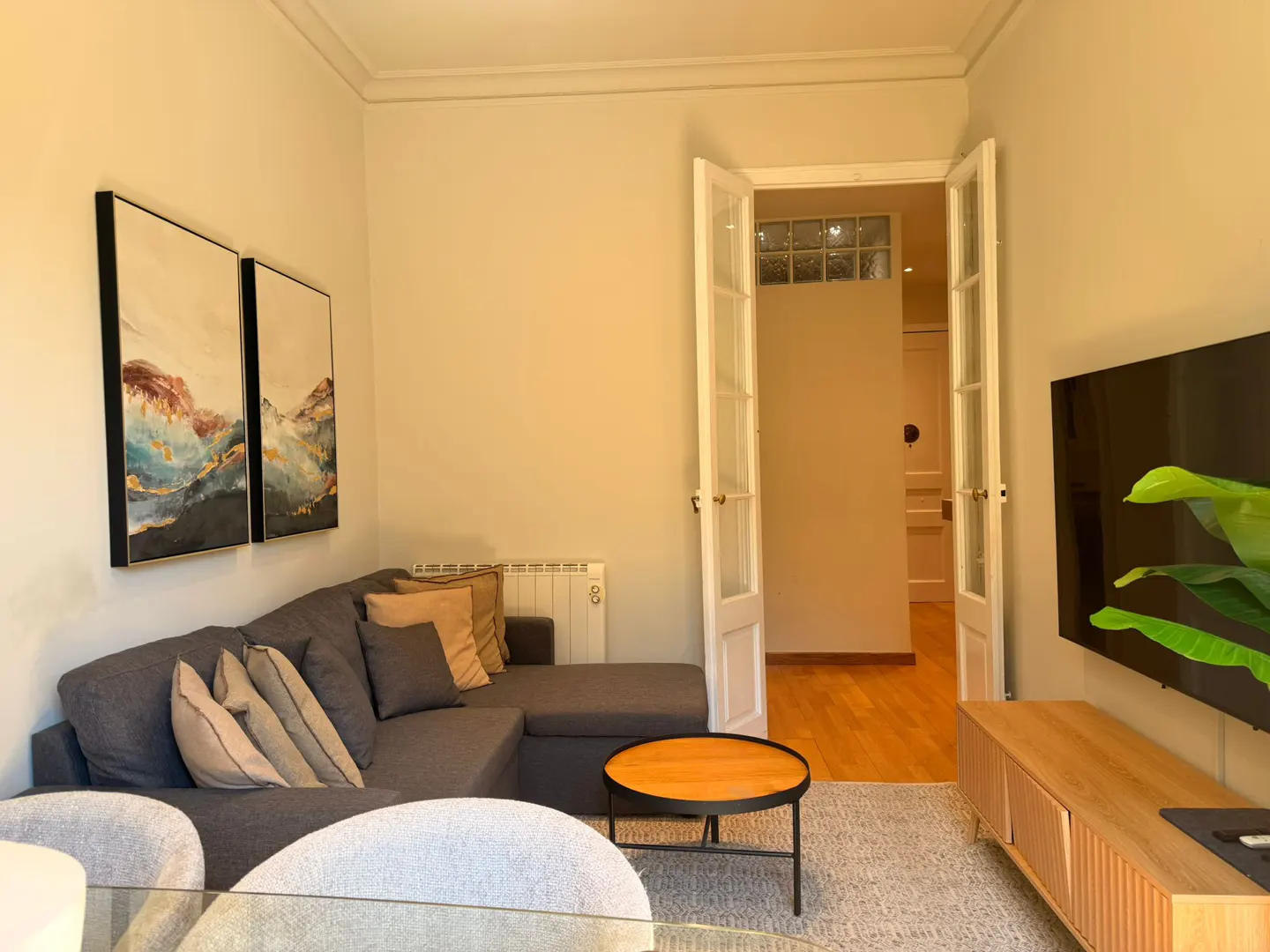 Living room with gray sectional sofa, abstract art, and round wood table. Open French doors lead to a hallway with wood floors.