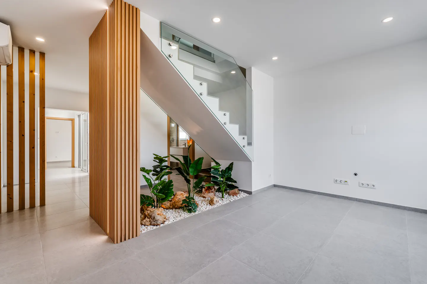 Bright, modern interior with gray tile floor, white walls, and wooden slat dividers. A white staircase with glass railing sits above a small indoor garden.