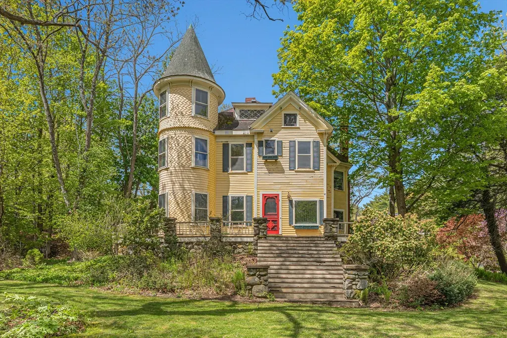 Yellow Victorian house with a turret, red door, and stone steps, surrounded by green trees under a blue sky.