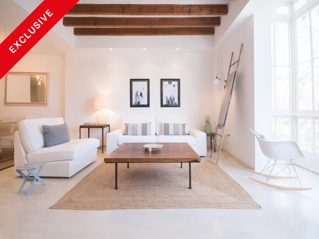 Bright living room with white walls, wooden ceiling beams, and a jute rug. White sofas, a wooden coffee table, and a rocking chair complete the scene.