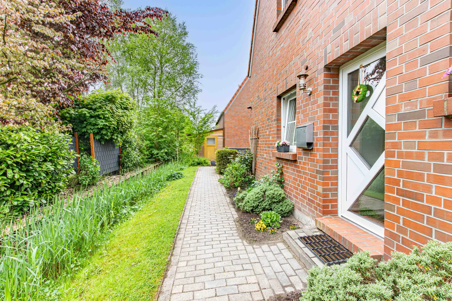 Brick house exterior with a white door, green wreath, and a stone walkway lined with lush greenery.