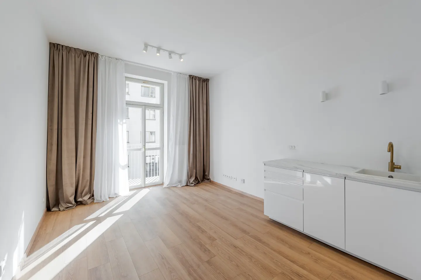 Bright, empty apartment with wood floors, white walls, and a kitchen counter. Curtains frame a window with a view of a building.