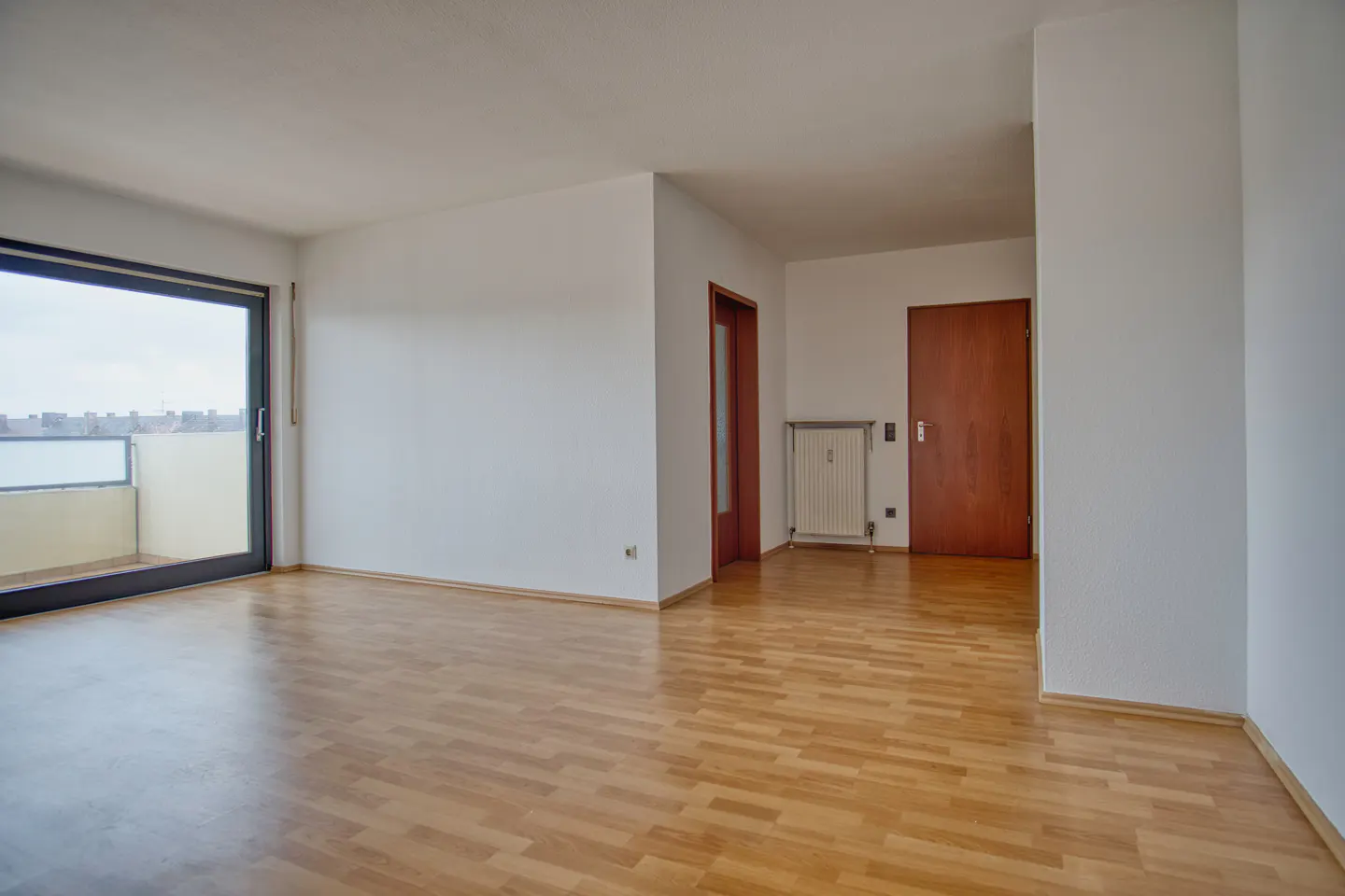 Empty room with light wood floors, white walls, and a sliding glass door to a balcony. Two brown doors and a radiator are visible.