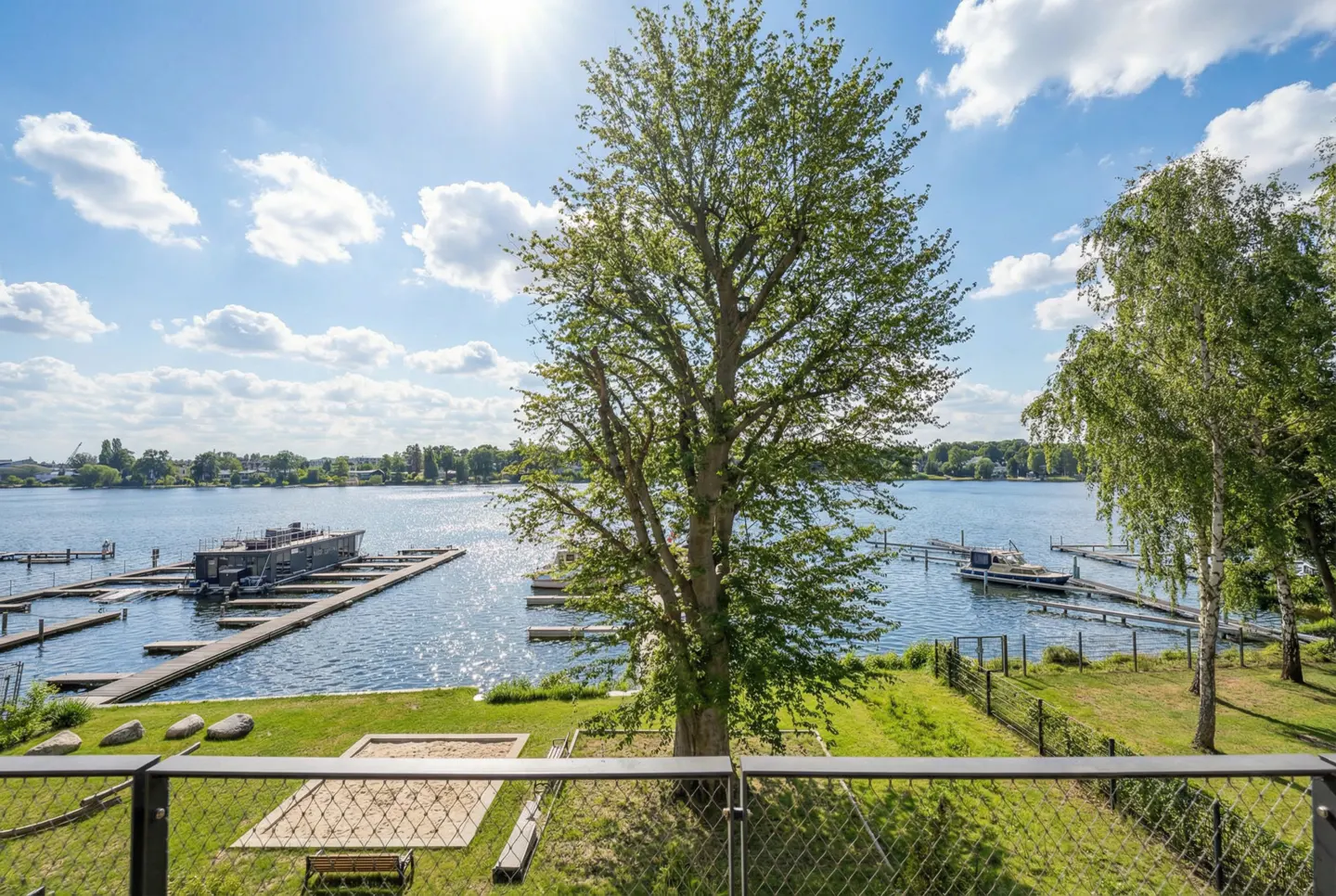 View of a lake with docks and boats, seen through a metal fence with a tree in the foreground, under a blue sky with clouds.