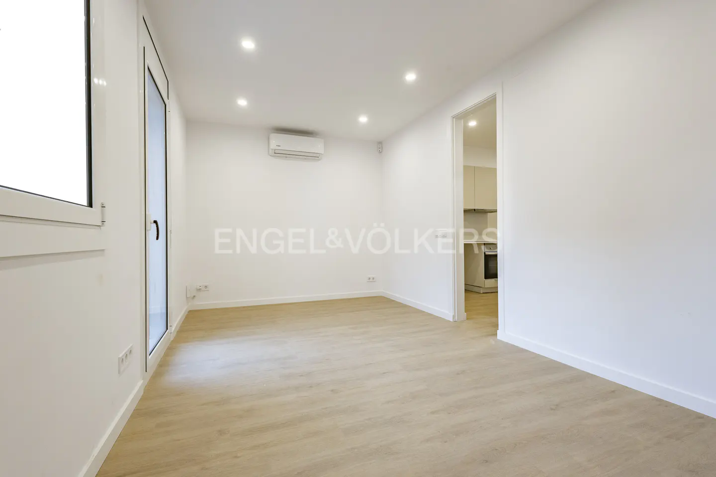 Bright, empty room with light wood floors and white walls. A doorway leads to a kitchen. Engel & Völkers logo on the wall.