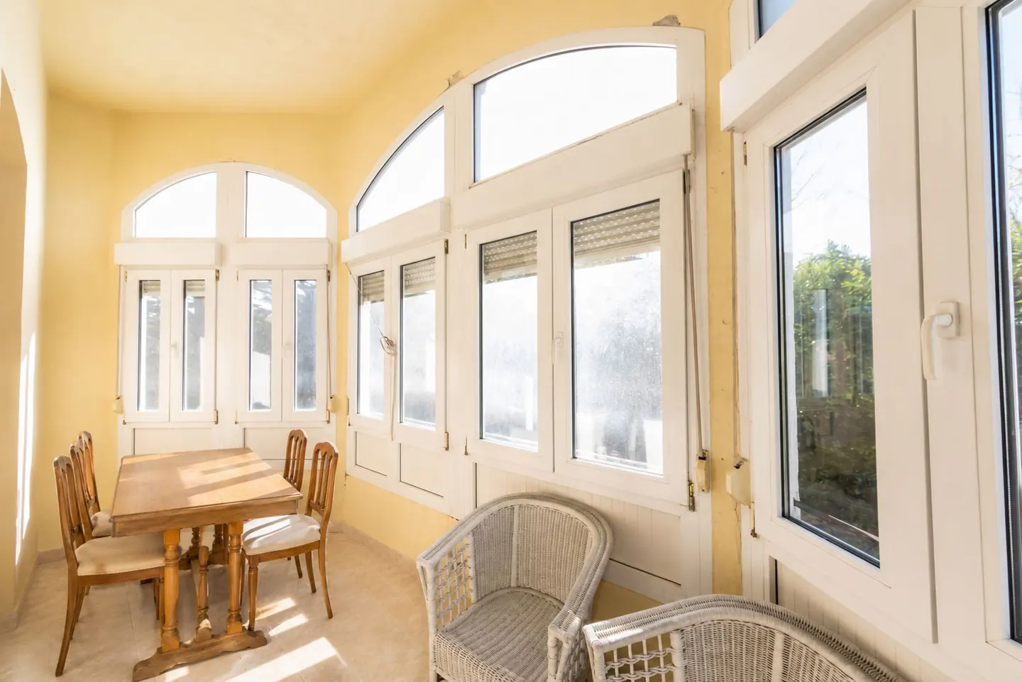Bright sunroom with yellow walls, white trim, and large windows. A wooden table and chairs sit near wicker armchairs.